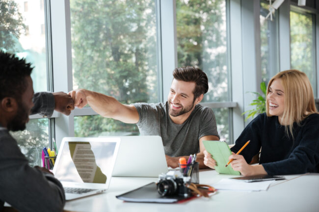 Photo of smiling young colleagues sitting in office coworking using laptop computers and tablet. Looking aside.